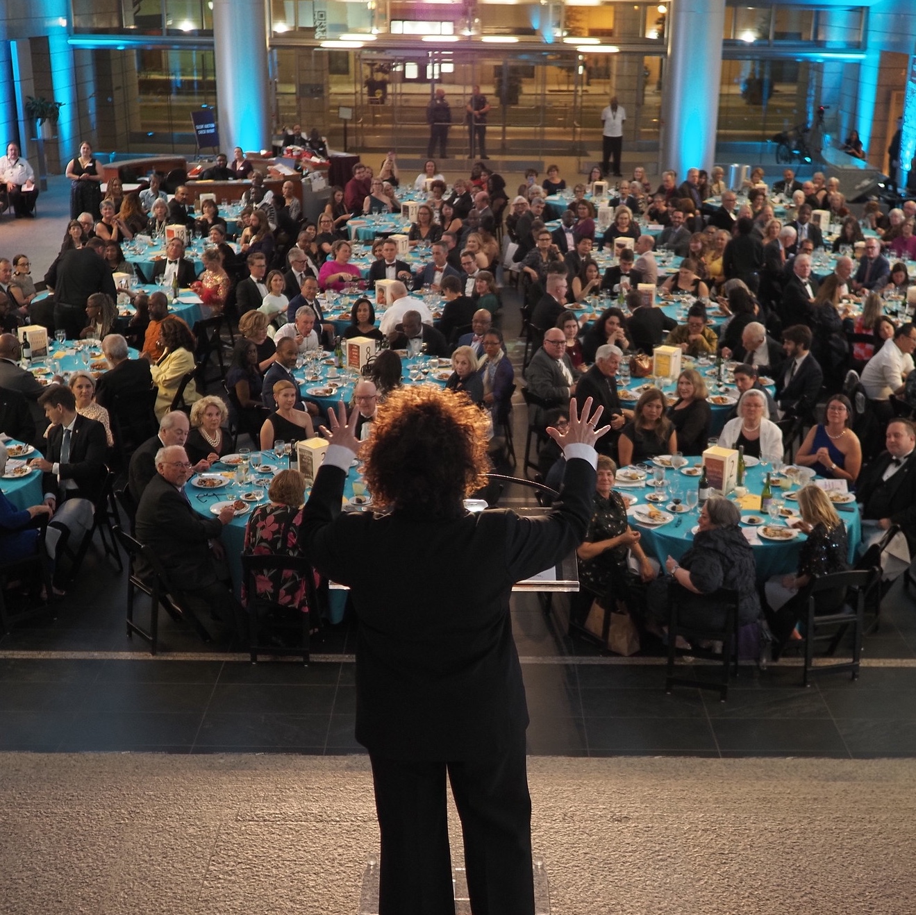 A woman standing on a elevated platform in front of a crowd sitting at tables during an event.