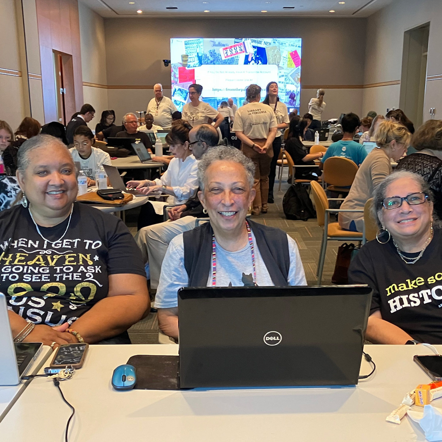 Three women face the camera in a crowded room. They are sitting at a table, with a laptop in front of the middle person. 