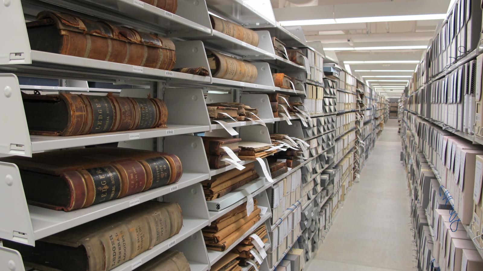 Image shows volumes and boxes on library shelving down a long aisle.