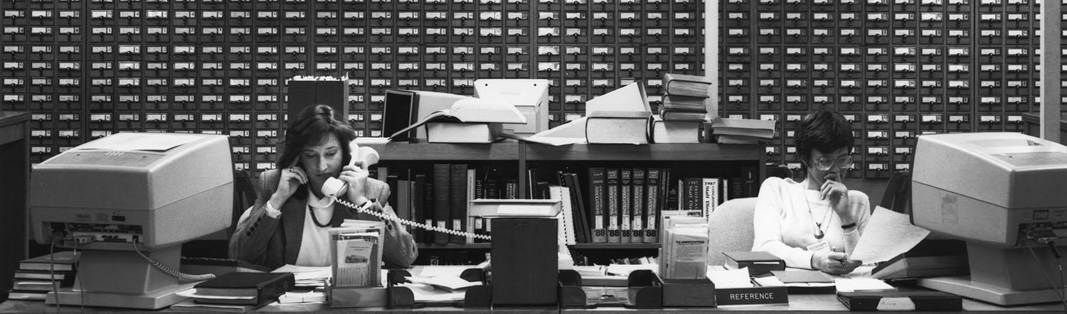 Two women sitting at a long desk, with computer terminals next to them. One is on the phone, while the other reads a piece of paper. Behind them is a large card catalog.