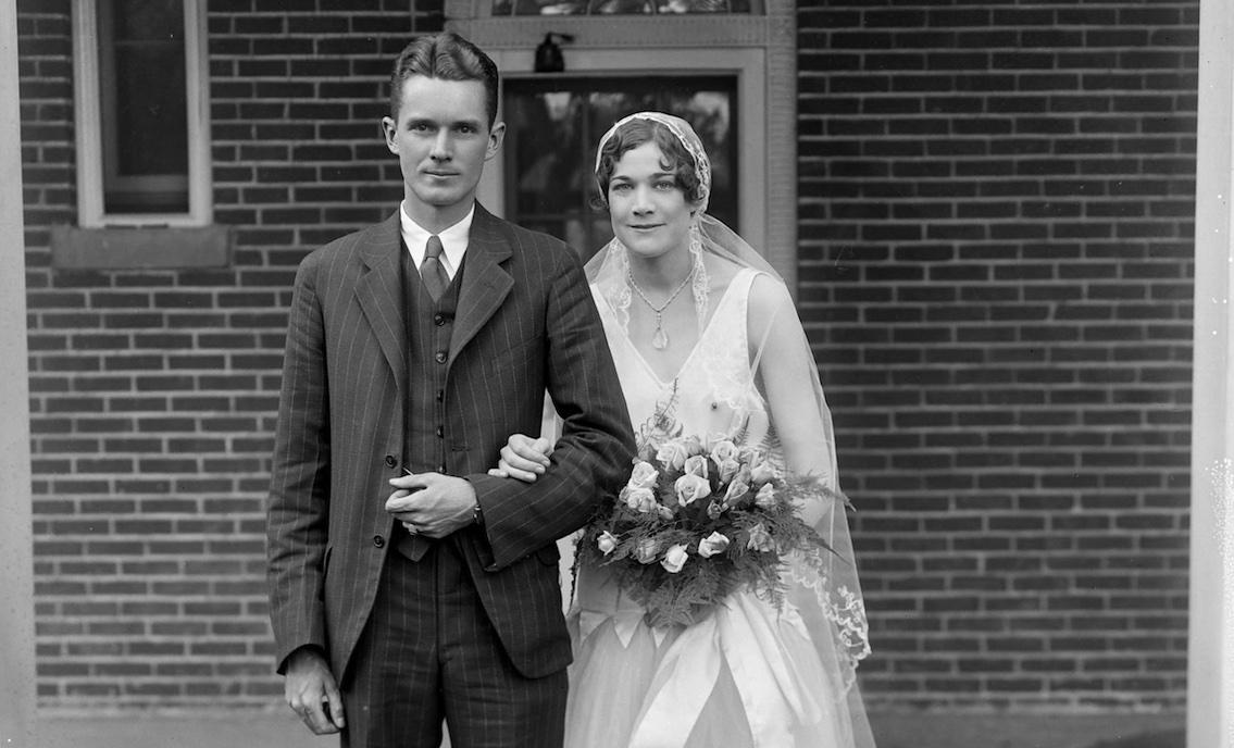 A wedding photograph featuring a man in the three-piece suit and a bride in sleeveless white dress to below her knees. She wears white stockings and shoes and a veil. She carries a bouquet of light colored roses.