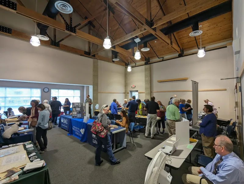 Large room filled the people moving around between tables. The tables have material and resources for the archive manning the table.