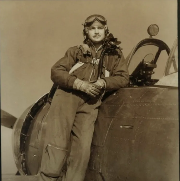 Image shows a man in the 1940s dressed in a pilot's uniform with goggles on his forehead and an oxygen mask on his shoulder. He is standing on the wing of his plane, leaning against the fuselage where the cockpit is visbible.