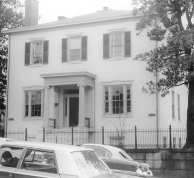 Image shows a large white mansion built in the 1840s. It has black shutters on three windows on the second floor, a covered portico in the middle of the first floor at the entrance, which is flanked by a window on each side. There is a slight iron fencing around the property and there are cars circa 1960s in front of the house.