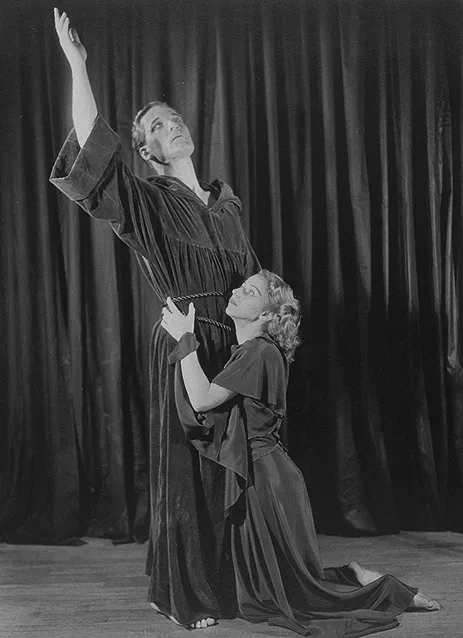 A man and woman dressed in robes against a curtain backdrop. The man is standing with one arm raised to the sky while the woman kneels on the ground, clasping the man at his waist.