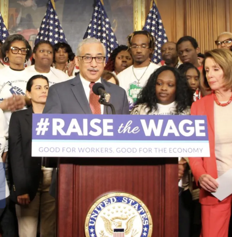 A man, Congressman Bobby Scott, dressed in a suit with a red tie, stands at a podium that shows part of the great seal of the United States and a sign that says "#Raise the Wage good for workers good for the economy." There are three rows of people behind him as he speaks. To his left is Nancy Pelosi, former representative from California. There are partially viewable American flags, showing only the blue fields with white stars.