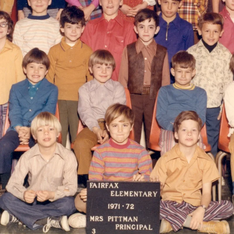 The image is a sample from a school class picture. Three boys sit cross-legged in the front, holding a sign that says Fairfax Elementary 1971-72 Mrs. Pittman Principal" with a "3" in the bottom left, possibly indicating the students being in 3rd grade. There are 3 male students seated behind them, and there are more students standing behind that row.