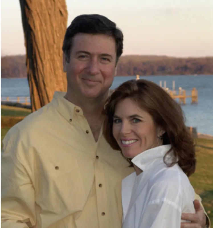 A photograph of George and Susan Allen standing closely together in front of a body of water. He is wearing a yellow long sleeve dress shirt and she is wearing a white shirt with an upturned collar. There is a tree and a small dock behind them. In the distance there are trees without leaves. 