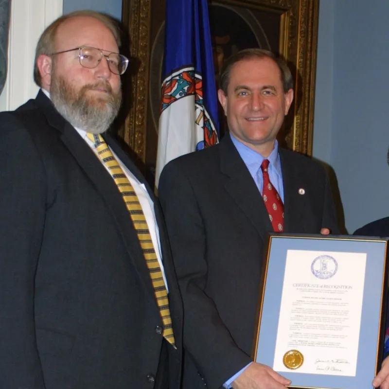 Photograph of two men standing. One is holding a framed document with a blue mat and a gold seal.