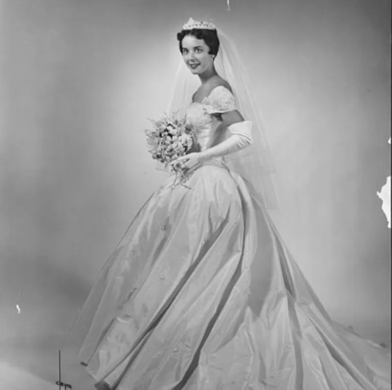 A picture of a woman in a wedding gown. She is wearing a tiara veil and is holding a bouquet of flowers. The photo is black and white and dated from 1956.