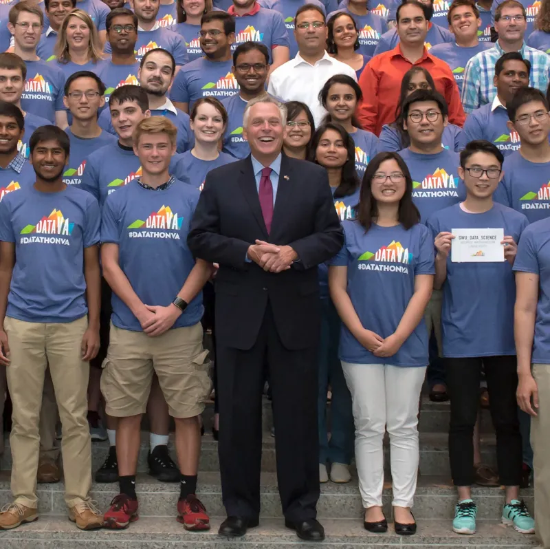 Virginia Governor Terry McAuliffe, dressed in a dark suit, stands in a photograph of a number of people dressed in blue tshirts that say DataVA #datathonVA in 2017.