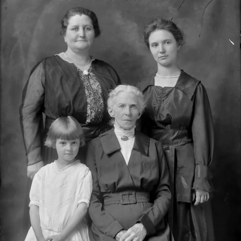 A studio portrait of several generations of women. The oldest woman, dressed in black, is seated, while the youngest, dressed white, leans on her right side. Behind them are two women standing, both dressed in black. 