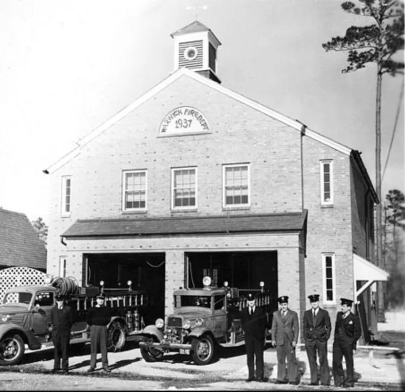 Image shows the Hilton Village fire house on Main Street at Warwick in 1937. There are several uniformed men standing in front of the building with two firetrucks with hoses and ladders on them.