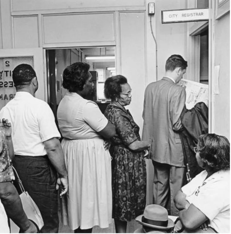 Image shows two men and two women in line near a window in an office that has a sign above it reading "City Registrar." The man at the front of the line by the window is reading a newspaper. There is a woman seated next to the window looking toward those in line.