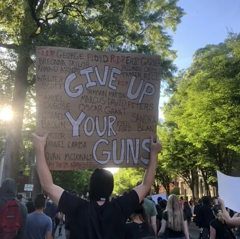 Image shows a protester holding up a hand-made sign that says "Give Up Your Guns" and includes names of Black individuals killed during interactions with police. There are other protestors in the image, that shows a blue and sunny sky and green trees.