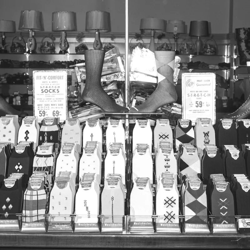 Image shows a display of men's socks with three rows of folded socks in various colors and patterns. There are two signs on either end advertising the various socks, such as Fit-'N'-Comfort boys' and men's cotton and nylon stretch socks and men's big and fancy stretch socks. Behnd the display, there are shelves with lamps and other homegoods for sale.