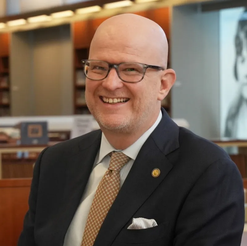 Photograph of a man smiling. He has glasses and is wearing a blazer with a white shirt and tie.