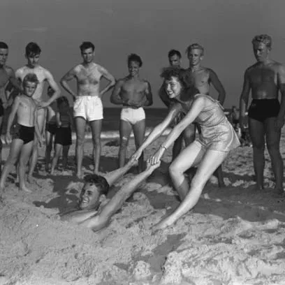 Image is on a beach by the ocean. A woman stands in the sand, pulling a man by the arms, who is buried in the sand. They are surrounded by several males dressed in swim trunks.