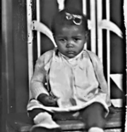 Image shows an infant dressed in a white dress and a sweater sitting on a chair.