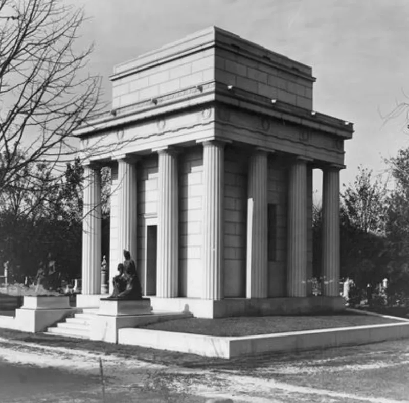 View of the John H. Core Mausoleum in Elwood Cemetery, Norfolk, Va. The mausoleum is in a classicizing architectural style, with Egyptian styled architectural elements. Columns run around the exterior, surrounding a central building. On the stairs are two matching bronze sculptures, both with two women, clothed and not clothed. One is seated, wearing an Egyptian headress with another woman seated beside the first. Each sculpture is slightly different, with the seated woman holding different attributes and a