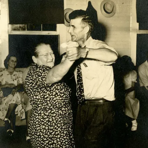 Image shows an older couple dancing. The woman is wearing a patterned dress and the man is in dress pants and a short-sleeved shirt with a tie. There are people sitting in the background, enjoying themselves.