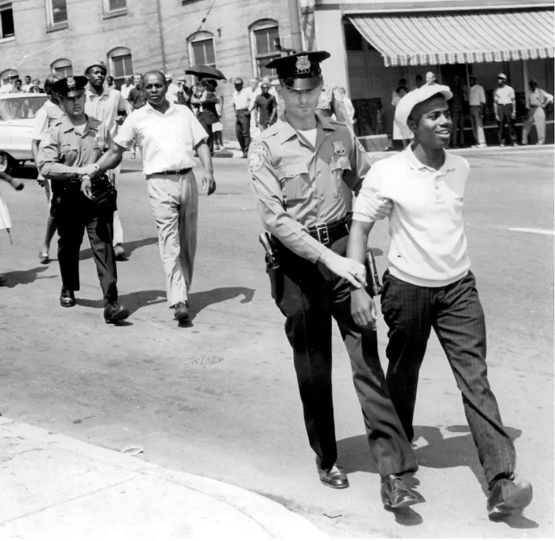 An image of two Black men being led by two white police officers on a city street. There is a crowd in the background watching the events taking place.