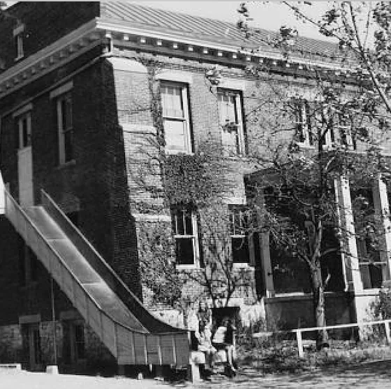 Black and white image shows a brick two story building with a slide attached to the side, running from a second floor window to the ground. There are two children at the bottom of the slide.