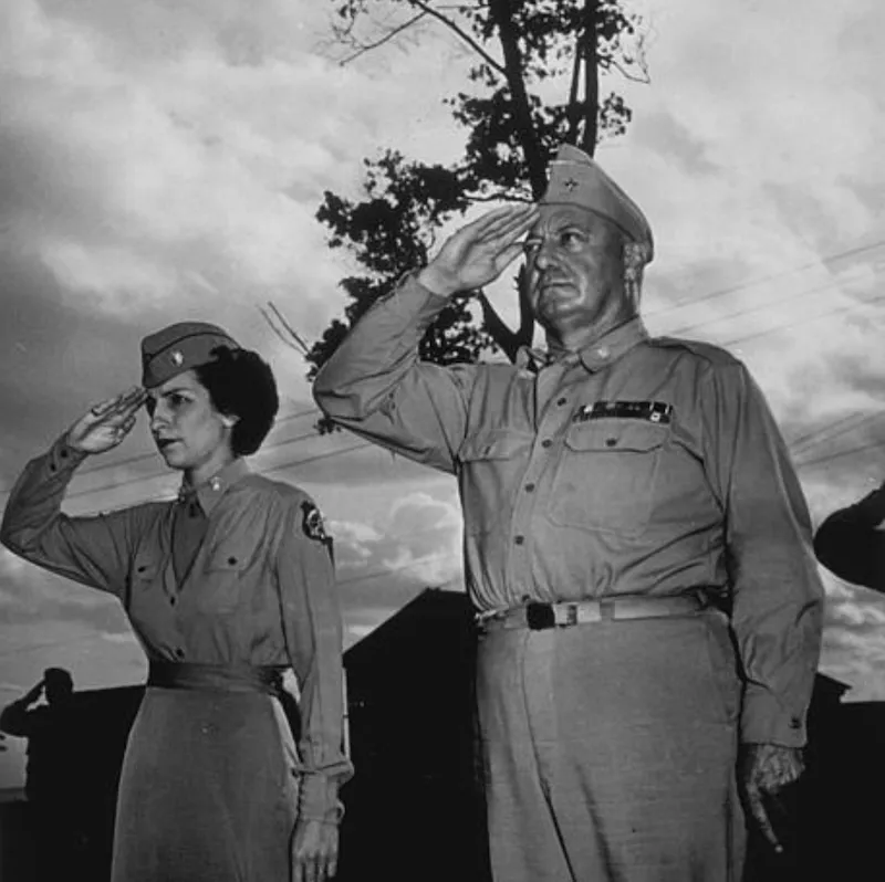 A woman and a man, both wearing Army uniforms, stand at attention, saluting. Behind them is a cloudy sky and a single tree. Faintly seen in the back, shadowed in black, is the outline of a building and another soldier saluting.