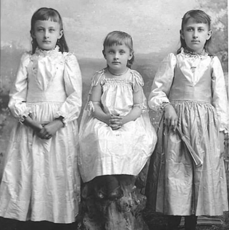 This studio photograph shows three children, each dressed in light colored dresses, likely dated in the late 19th or early 20th century. The smallest child is sitting on a tree stump, flanked by the others. The background evokes trees, a field, and the sky.