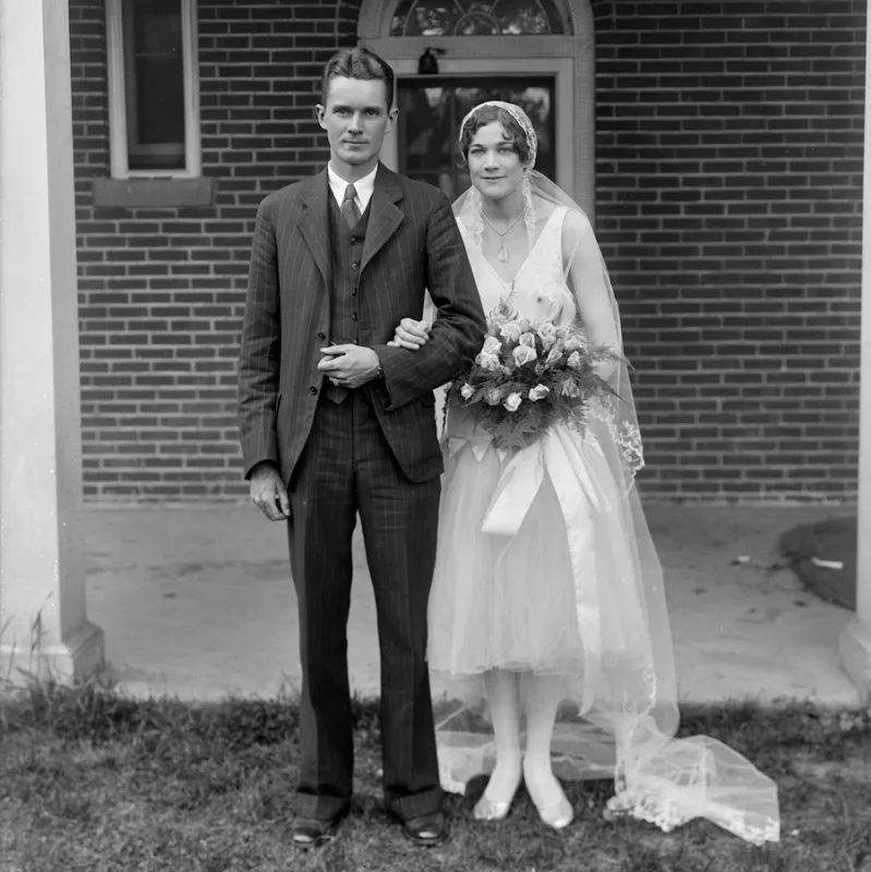A wedding photograph featuring a man in the three-piece suit and a bride in sleeveless white dress to below her knees. She wears white stockings and shoes and a veil. She carries a bouquet of light colored roses.