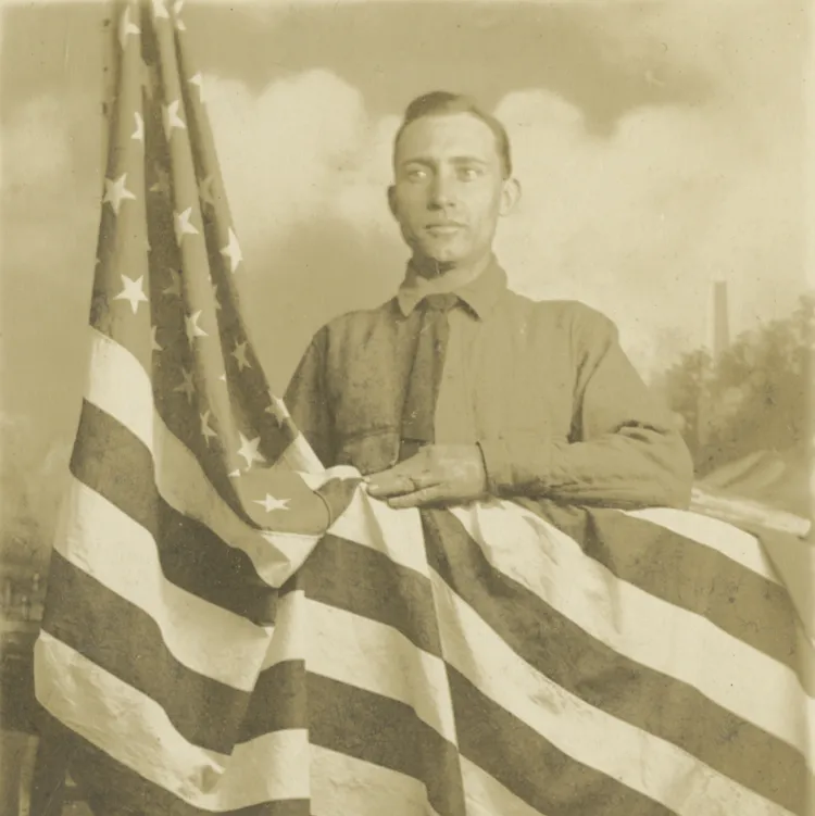 A World War I era soldier dressed in a long-sleeved shirt and tie poses behind a draped American flag, with his arm resting upon the flag.